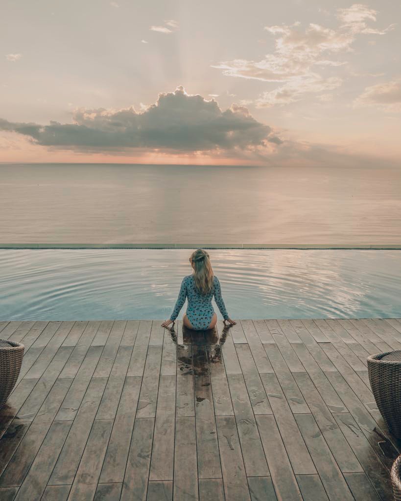 A woman sitting on the edge of an infinity pool with a breathtaking ocean view at sunset.